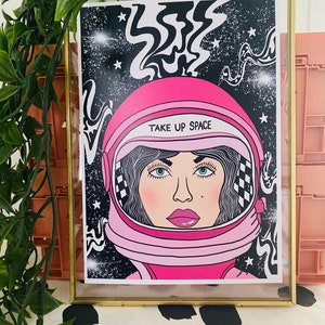 May include: A pink astronaut helmet with a black and white checkered pattern on the side. The helmet has a white visor with the text "TAKE UP SPACE" printed on it. The helmet is set against a black background with white stars and swirling lines.