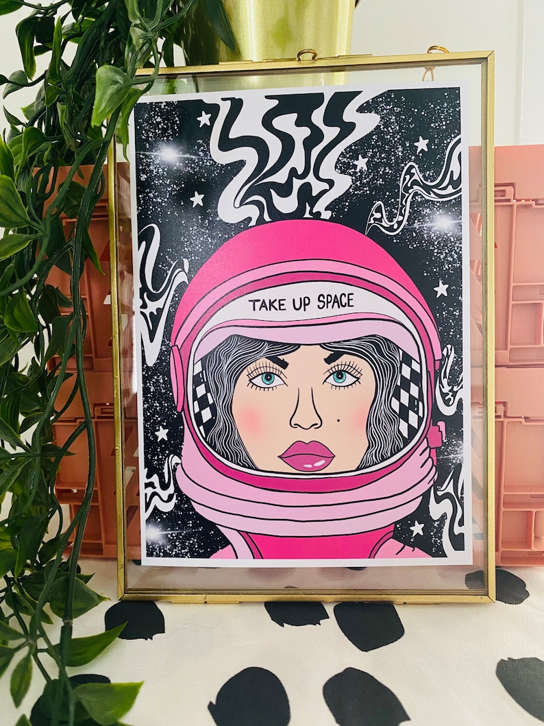 May include: A pink astronaut helmet with a black and white checkered pattern on the side. The helmet has a white visor with the text "TAKE UP SPACE" printed on it. The helmet is set against a black background with white stars and swirling lines.