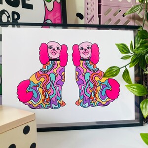 May include: A framed print of two cartoon dogs with pink fur and psychedelic patterns. The dogs are wearing collars with charms. The print is on white paper with a black frame.