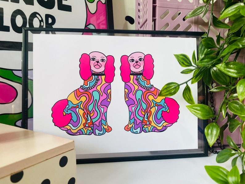 May include: A framed print of two cartoon dogs with pink fur and psychedelic patterns. The dogs are wearing collars with charms. The print is on white paper with a black frame.