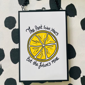 K&ouml;nnte beinhalten: Ein schwarzer gerahmter Druck mit wei&szlig;em Hintergrund und einer Illustration einer gelben Zitronenscheibe. Der Text lautet "The past was yours but the future's mine."