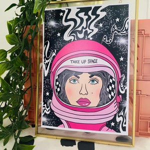 May include: A pink astronaut helmet with a black and white checkered pattern and the text "Take Up Space" on a black background with white stars and swirling lines.