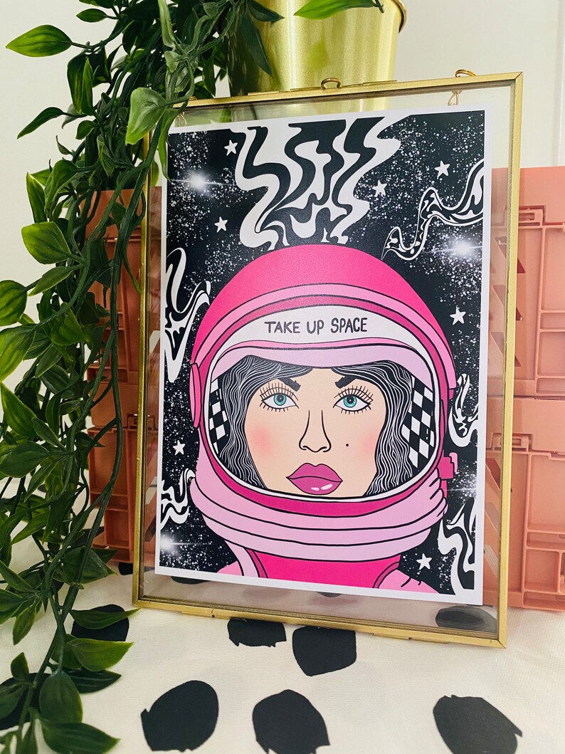 May include: A pink astronaut helmet with a black and white checkered pattern and the text "Take Up Space" on a black background with white stars and swirling lines.