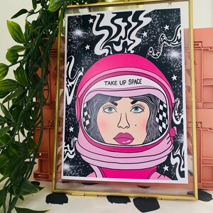 May include: A framed print of a woman wearing a pink astronaut helmet with the text "TAKE UP SPACE" on the helmet. The background is black with white stars and swirls.