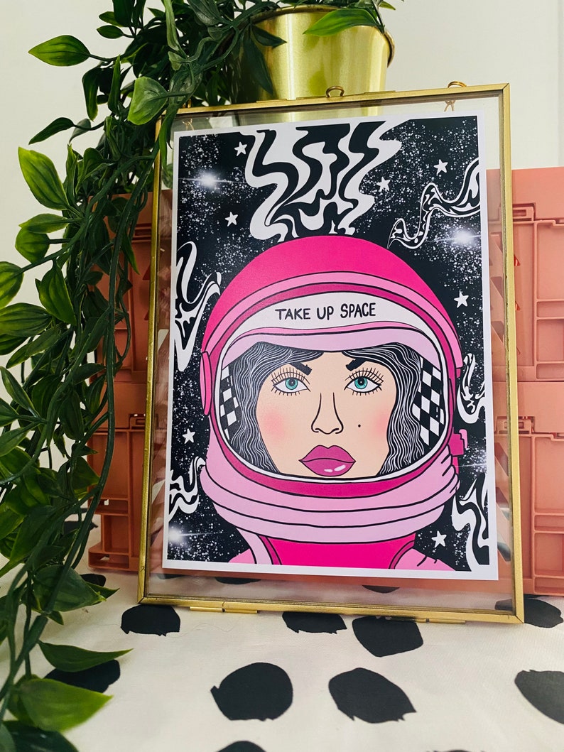 May include: A framed print of a woman wearing a pink astronaut helmet with the text "TAKE UP SPACE" on the helmet. The background is black with white stars and swirls.
