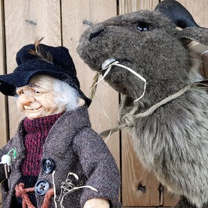May include: A brown and gray plush gnome with a black hat and a white beard is holding a rope. The gnome is standing next to a brown and gray plush goat carrying a bag and a wooden ladder.