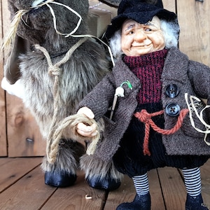May include: A whimsical gnome doll wearing a brown coat, black hat, and red scarf, holding a rope attached to a gray and brown plush donkey doll carrying a small package.