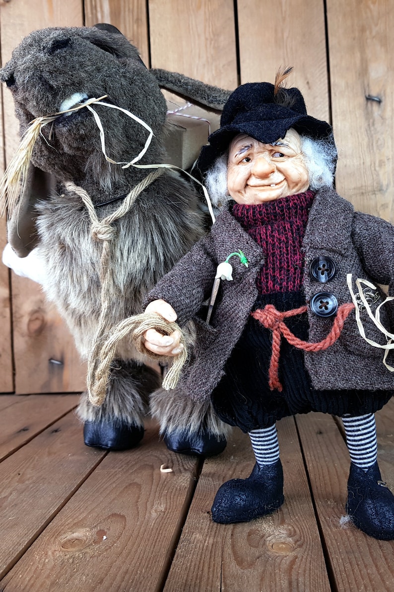 May include: A whimsical gnome doll wearing a brown coat, black hat, and red scarf, holding a rope attached to a gray and brown plush donkey doll carrying a small package.