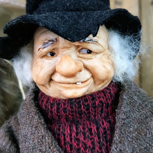 May include: A whimsical figurine of an elderly man with a white beard, wearing a black hat, a red and black knitted scarf, and a brown coat. He has a mischievous smile and a missing tooth.