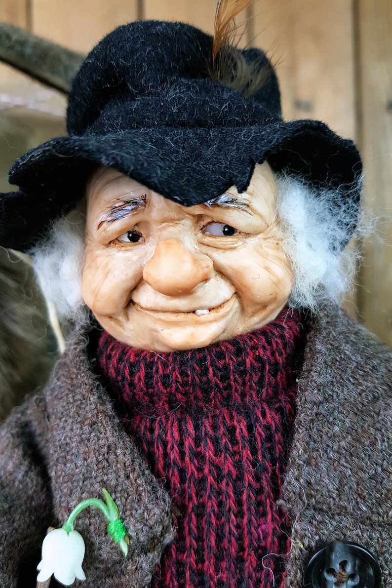 May include: A whimsical figurine of an elderly man with a white beard, wearing a black hat, a red and black knitted scarf, and a brown coat. He has a mischievous smile and a missing tooth.