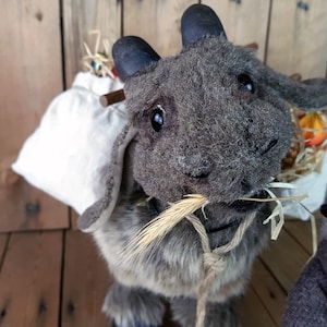 May include: A gray plush toy goat with a white sack on its back. The goat is holding a stalk of wheat in its mouth and has a rope tied around its neck.