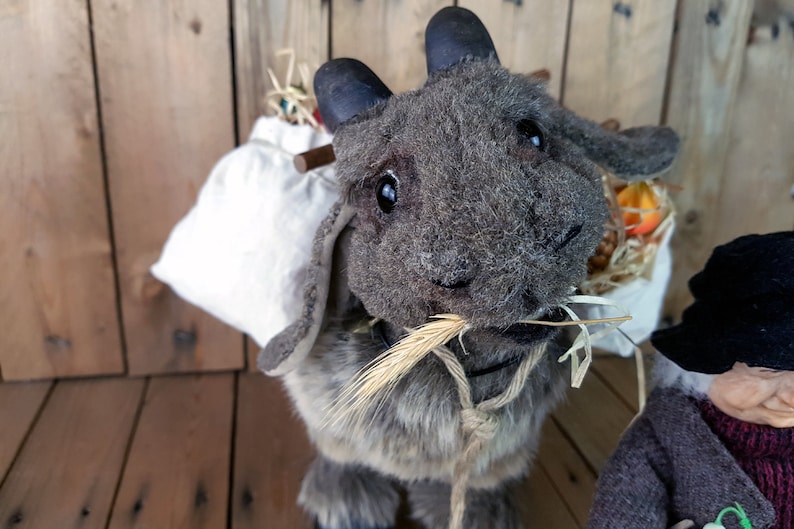 May include: A gray plush toy goat with a white sack on its back. The goat is holding a stalk of wheat in its mouth and has a rope tied around its neck.