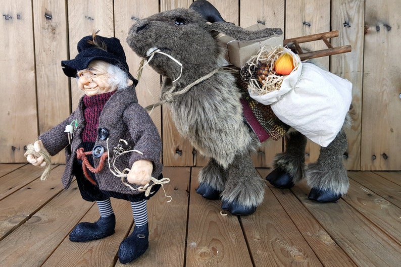 May include: A brown and gray plush gnome with a black hat and a white beard is holding a rope. The gnome is standing next to a brown and gray plush goat carrying a bag and a wooden ladder.