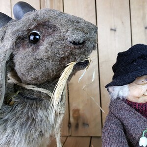 May include: A gray and brown stuffed goat with large horns and a long, thin piece of straw in its mouth. The goat is wearing a black collar and is standing next to a small, gray-haired, elderly doll wearing a black hat and a brown coat.