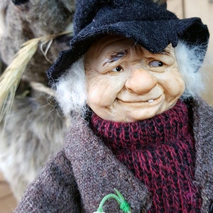 May include: A whimsical figurine of an elderly person with white hair and a black hat, wearing a brown coat with a red scarf and a white flower pinned to it. The figurine has a friendly expression and is holding a small, furry animal.