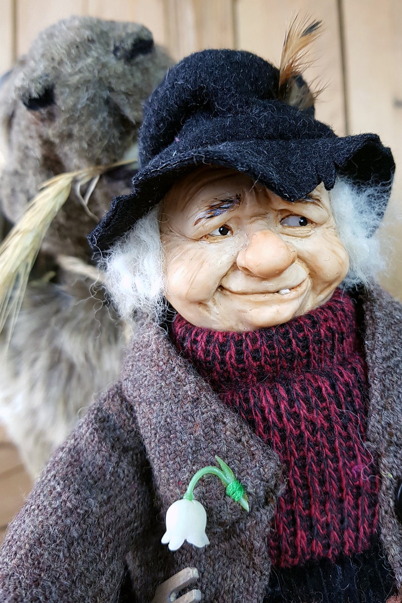 May include: A whimsical figurine of an elderly person with white hair and a black hat, wearing a brown coat with a red scarf and a white flower pinned to it. The figurine has a friendly expression and is holding a small, furry animal.