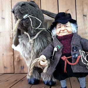 May include: A brown and gray plush donkey with a long, floppy ear and a brown rope tied around its neck. A small, gray-haired gnome with a black hat and a brown coat is holding the rope and smiling.