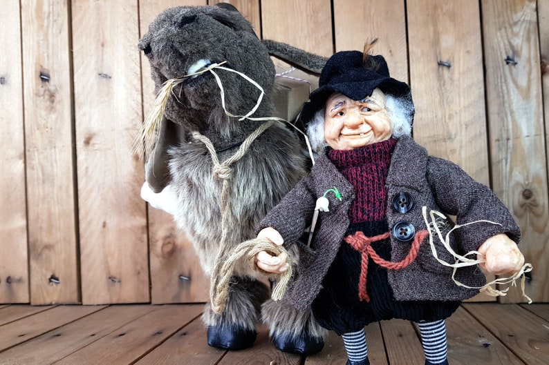 May include: A brown and gray plush donkey with a long, floppy ear and a brown rope tied around its neck. A small, gray-haired gnome with a black hat and a brown coat is holding the rope and smiling.