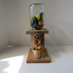 May include: A wooden gumball machine with a glass jar filled with colorful gumballs. The machine has a crank and a small wooden bucket underneath to catch the gumballs.