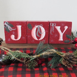 May include: Three red wooden blocks arranged to spell JOY, each featuring hand-painted white letters, grey bows, and green holly leaves. The blocks sit on a wooden base, accented with pine branches, pinecones, and dark metal bells.