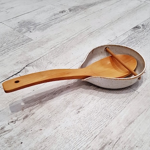 May include: White ceramic spoon rest with a wooden spoon resting inside. The spoon rest has a speckled glaze and a rough texture.