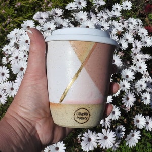 May include: A ceramic travel mug with a white lid, featuring a geometric design in white, peach, and gold. The mug has a speckled beige base and the text "Lillypilly Pottery" on the bottom. The mug is held in front of a background of white flowers.
