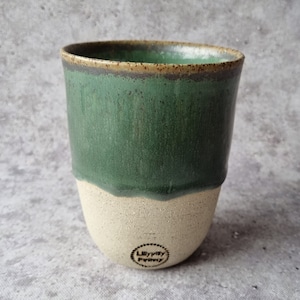 Könnte beinhalten: Eine grüne und weiße Keramiktasse mit braunem Rand. Die Tasse hat eine gesprenkelte Glasur und ein geprägtes Logo mit der Aufschrift "Lillylily Pottery".