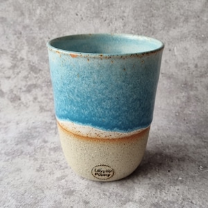 May include: A handmade ceramic tumbler with a two-tone design. The top half is a vibrant turquoise, transitioning to a sandy beige at the base. The rim has a rustic, speckled texture. The bottom of the tumbler has a small logo.