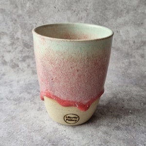 May include: A ceramic mug with a light green to pink ombre glaze. The mug has a drip glaze effect and a small logo on the bottom that reads "Lilly Mill Pottery".