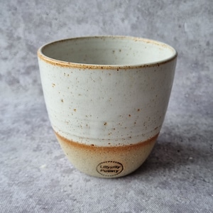 Könnte beinhalten: Weiße Keramiktasse mit braunem, gesprenkeltem Überzug und einem braunen Band am Boden. Die Tasse hat einen schwarzen Stempel auf dem Boden, auf dem "LillyPilly Pottery" steht.