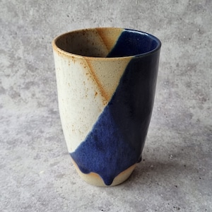 Puede incluir: Una taza de cerámica con un esmalte blanco, tostado y azul. La taza tiene un diseño abstracto único con un efecto de goteo.
