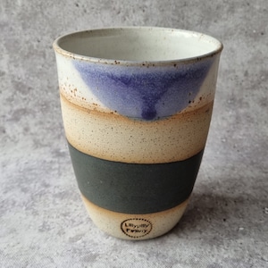 May include: A tall, ceramic mug with a striped design. The mug has a light brown base, a dark brown stripe, a light brown stripe, a dark gray stripe, and a light brown stripe. The top of the mug has a light blue and white design. The mug has a brown stamp on the bottom that says "Lilly pilly Pottery".