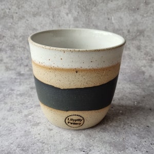 May include: A white ceramic mug with a black stripe and a beige stripe. The mug has a speckled glaze and a brown stamp that reads "Lilly pilly Pottery".