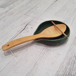 May include: A green ceramic spoon rest with a wooden spoon resting inside. The spoon rest has a textured, speckled finish.