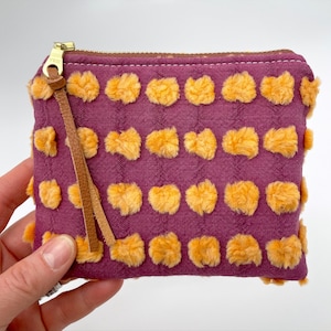 Peut inclure: Une petite pochette zippée violette avec un pompon en cuir marron. La pochette est décorée d'un motif de cercles jaunes flous.