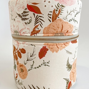 Op de afbeelding: Een witte cilindrische opbergcontainer met een ritssluiting. De container is bedekt met een bloemenpatroon met roze, oranje en groene bloemen en bladeren.