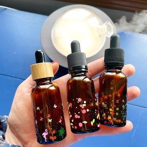 Puede incluir: Tres pequeños frascos de vidrio marrón con goteros, cada uno decorado con purpurina colorida y pequeñas pegatinas. Los frascos se sostienen en una mano sobre un fondo azul.