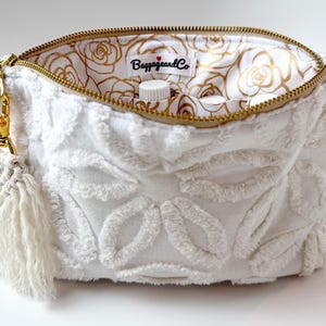 May include: A white, textured cosmetic bag with a gold zip and tassel. The interior lining features a gold rose pattern. The bag has a gold clasp and the label reads "BaggageandCo".