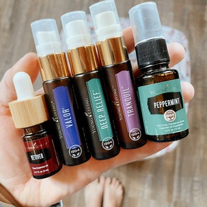May include: Five small bottles of essential oils, labeled "Valor", "Deep Relief", "Tranquil", "Peppermint", and "Vetiver". The bottles are all brown glass with gold caps and white labels. The labels have the Young Living logo and the name of the oil. The bottles are held in a hand.
