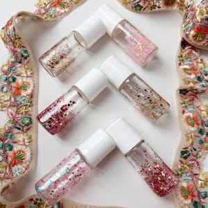May include: A collection of small, clear glass bottles with white caps, filled with colorful glitter. The glitter includes gold, pink, and red hues. The bottles are arranged on a white surface, with a decorative floral ribbon in the background.