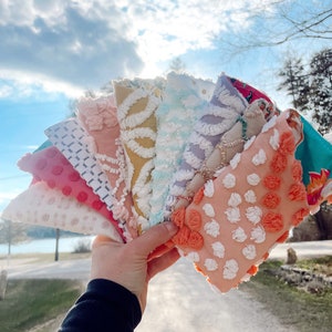 Op de afbeelding: Een hand houdt een waaier van kleurrijke, getextureerde stoffen vierkanten vast. De vierkanten tonen verschillende patronen en kleuren, waaronder roze met witte stippen, wit met een geometrisch ontwerp en perzik met witte en oranje plukjes. De achtergrond toont een zonnige hemel en een weg.