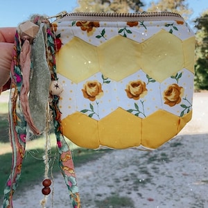 Könnte beinhalten: Kleine, handgefertigte Tasche mit gelb-weißem Blumenmuster. Die Tasche hat einen Reißverschluss und ist mit dekorativen Stoffquasten und Holzperlen verziert. Die Außenseite ist ein Patchwork aus gelben und weißen sechseckigen Formen mit Rosenmustern.