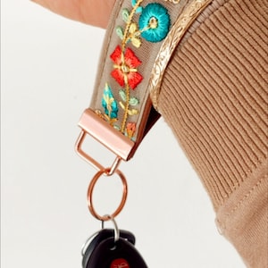 May include: A beige key chain with a floral embroidered strap and a rose gold key ring. The key ring has a black car key fob attached.