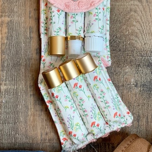 May include: A pink fabric pouch with a floral pattern and a butterfly charm. The pouch has six slots for holding essential oil roller bottles. Three gold roller bottles and three clear glass bottles are visible in the slots.