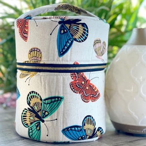 May include: A white canvas zippered pouch with a black zipper and a handle. The pouch is decorated with a colorful butterfly pattern. The pouch is sitting on a wooden surface next to a white diffuser.