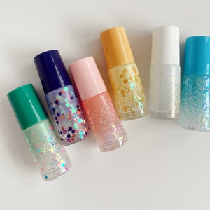 May include: Six small glass bottles with different colored lids and glitter inside. The bottles are arranged in a row on a white surface.