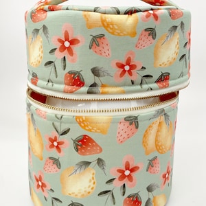 May include: A green fabric storage container with a zipper closure and a handle. The container has a floral pattern with lemons, strawberries, and flowers.