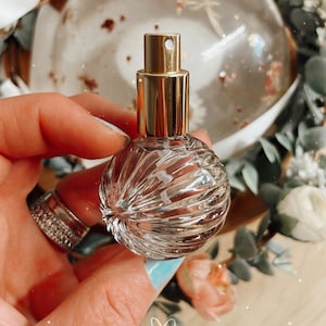 Può includere: Un flacone di profumo in vetro trasparente con un atomizzatore dorato e un design testurizzato e a coste. Il flacone è tenuto in una mano.