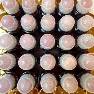 May include: A collection of twenty-five amber glass bottles arranged in a square grid, each topped with a white plastic collar and a smooth, spherical light pink rose quartz gemstone rollerball applicator, viewed from directly above.