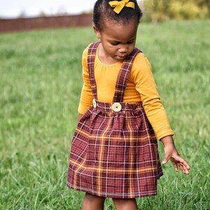 May include: A young girl wearing a yellow long-sleeved shirt with a plaid skirt and suspenders. The skirt has a brown, white, and orange plaid pattern. The girl is standing in a grassy field.